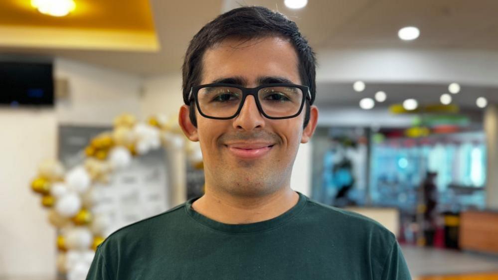 Dhillon Lalji looking at the camera, wearing glasses and a green top and smiling