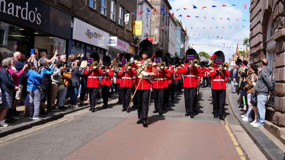 The guards marching through the street towards the camera. There is red and white bunting above them and people lining the street.
