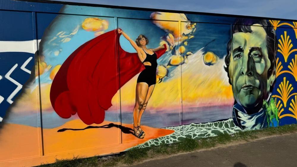 A painting on the hoarding of a woman in swimwear with a red cape catching the wind. She is standing on a sand dune with waves breaking on the shore. On the right of the image, Arthur Wellesley, Duke of Wellington - who's nickname was The Iron Duke.
