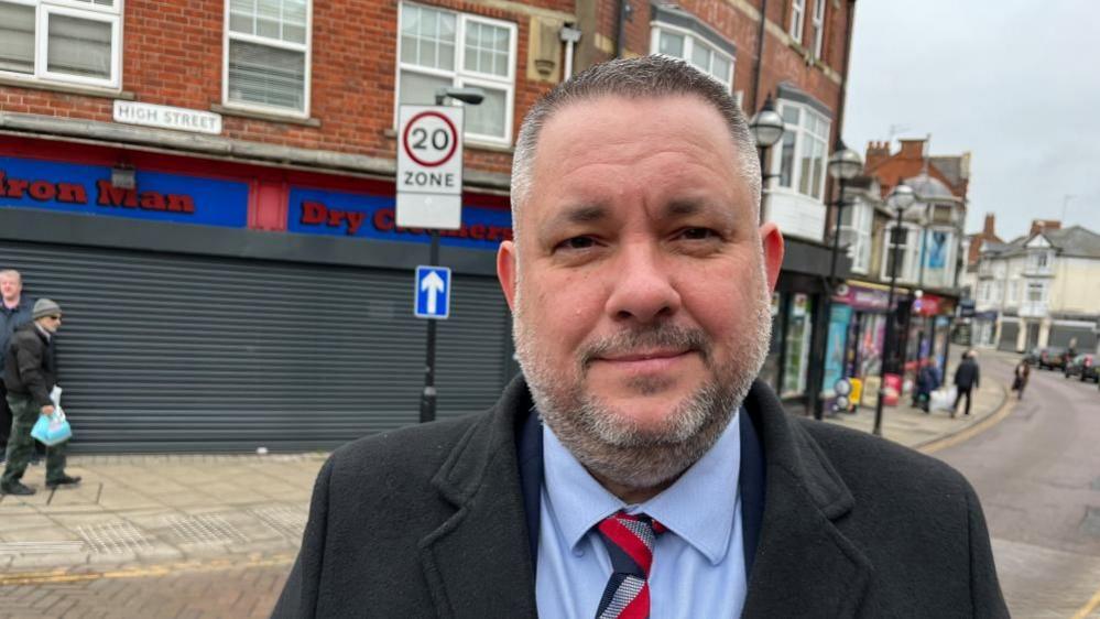 Jason Smithers stands on a high street, in a dark coat, blue shirt and striped tie