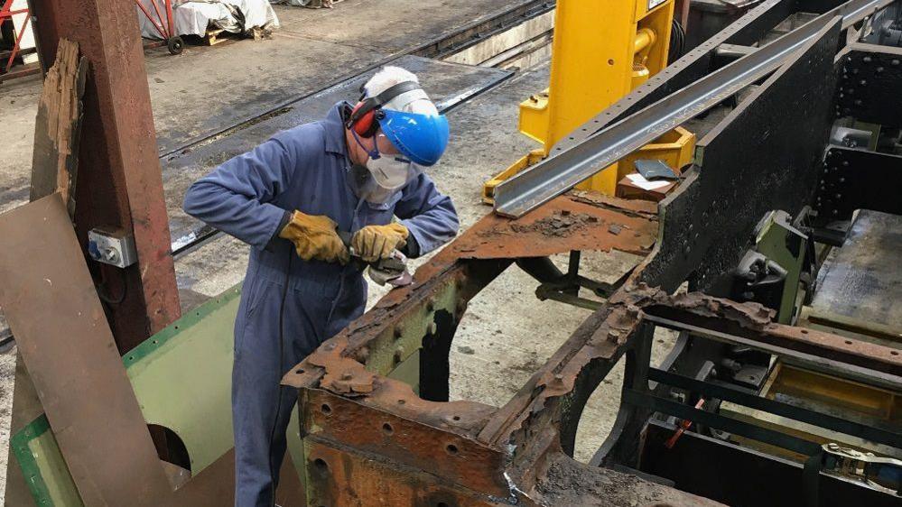 A man in a blue boiler suit, helmet and other protective gear using a power tool on the rusty locomotive in a workshop