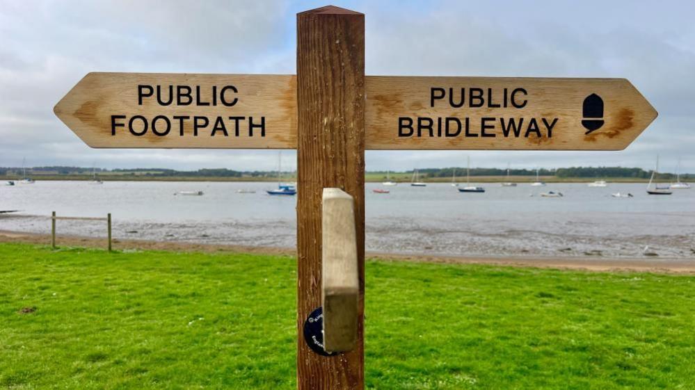 There is a three-way wooden sign post which has public footpath written on two arrows pointing in opposite directions. There is a symbol of an acorn in one direction. The symbol is for the England coast path. In the background is there Deben Estuary with bots bobbing around.
