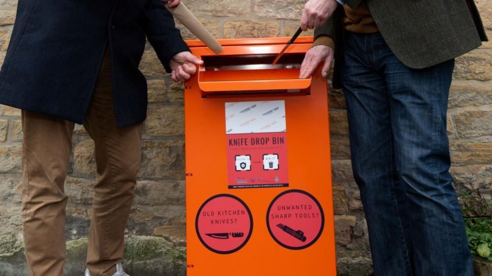 Two men stand on either side of an orange knife bin. Their torsos and heads and out of frame but they are each pretending to put a knife into the bin. 