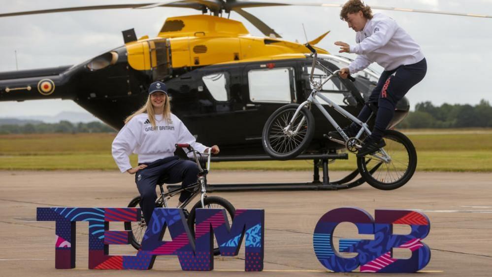 Charlotte and Kieran on BMX bikes in front of a helicopter and behind a TEAM GB sign