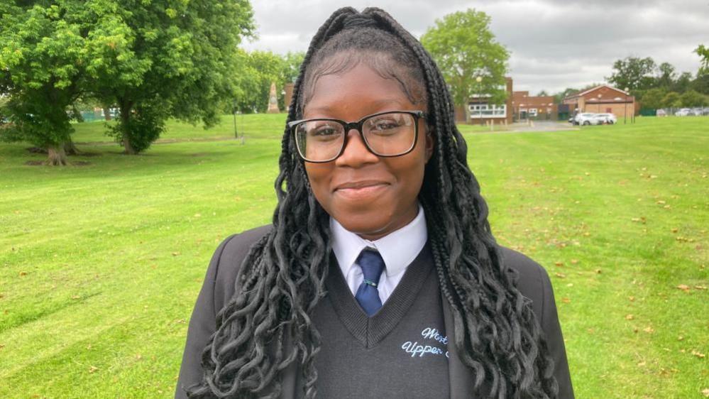 Franklina is photographed on a green field outside the school building, she is wearing glasses and a dark school uniform, with a blue tie