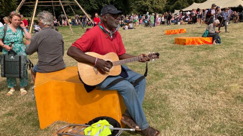 A man wearing a dark-coloured cap, red T-shirt and blue trousers sits on a seat covered with an orange blanket playing a guitar. A man wearing a grey top is also sitting on the seat talking to a woman in a green top. There is a swing behind them and a large crowd of people in front of a marquee in the background.