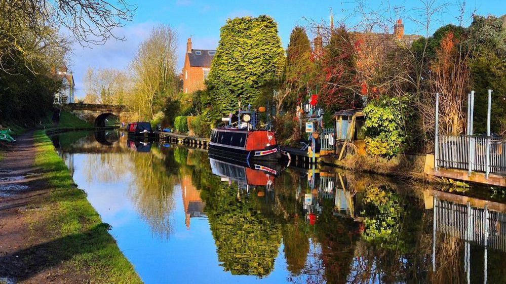 Two canal boats sit on water on a canal between two banks with trees behind them and a blue sky overhead, all reflected in the calm water.