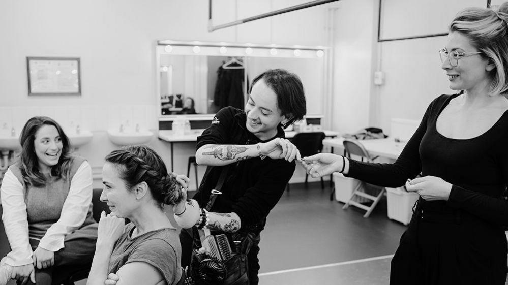 Two cast members and two hair and make-up artists laugh as they get ready. There are sinks and mirrored dressing tables behind them.