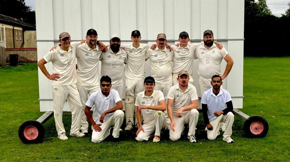 Eleven men in cricket whites standing and kneeling in a team line-up in from of a white sight screen