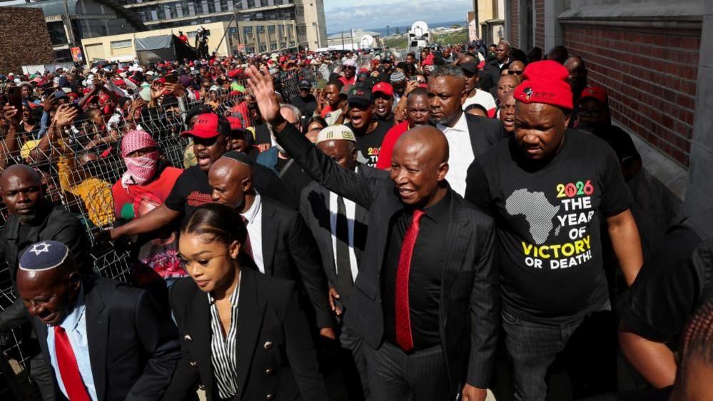 Julius Malema in a black suit and red tie walks out of court and waves at supporters who can be seen in the background.