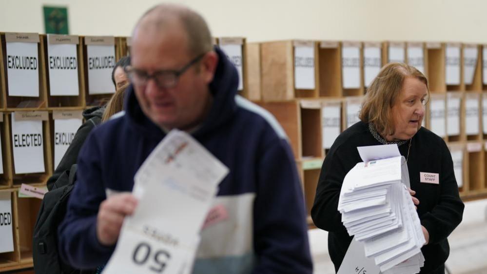 Two people seen at a count centre in the Republic of Ireland. The one in the foreground is blurred while the woman behind holding a stack of papers is the main focus