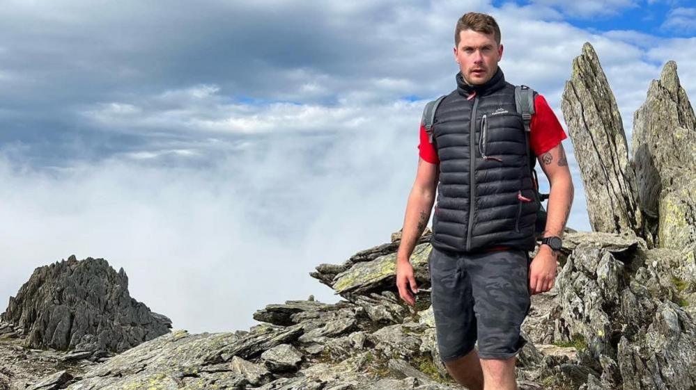 A dark haired man up a high mountain, wearing a red t-shirt under a blue padded body warmer and dark shorts, with jagged rocks in the background and a cloudy sky.
