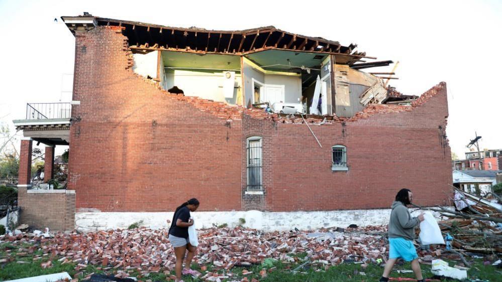 Bricks from a collapsed wall can be seen on the ground as two residents carry belongings away from a heavily damaged home in St Louis, Missouri, on 16 May 2025. The roof and several rooms on the upper floor are damaged and exposed.