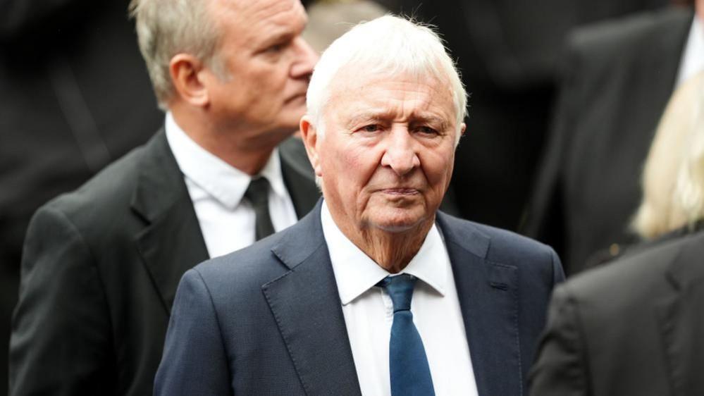 Mike Summerbee stands in a crowd wearing a navy suit and a serious expression.