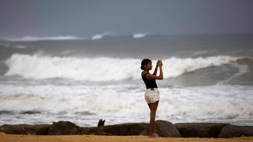 A woman wearing white shorts and a black tank top takes a photo of large waves as they break on the beach in the Dominican Republic.