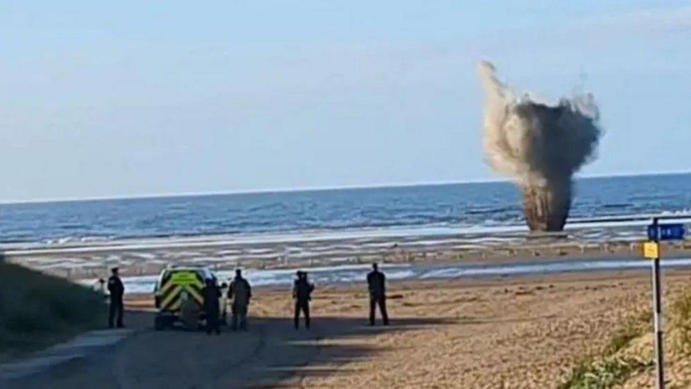 A picture of Ainsdale Beach as the WW2 bomb is exploded underwater. Smoke from the detonation can be see above the water, and officers are standing to watch.