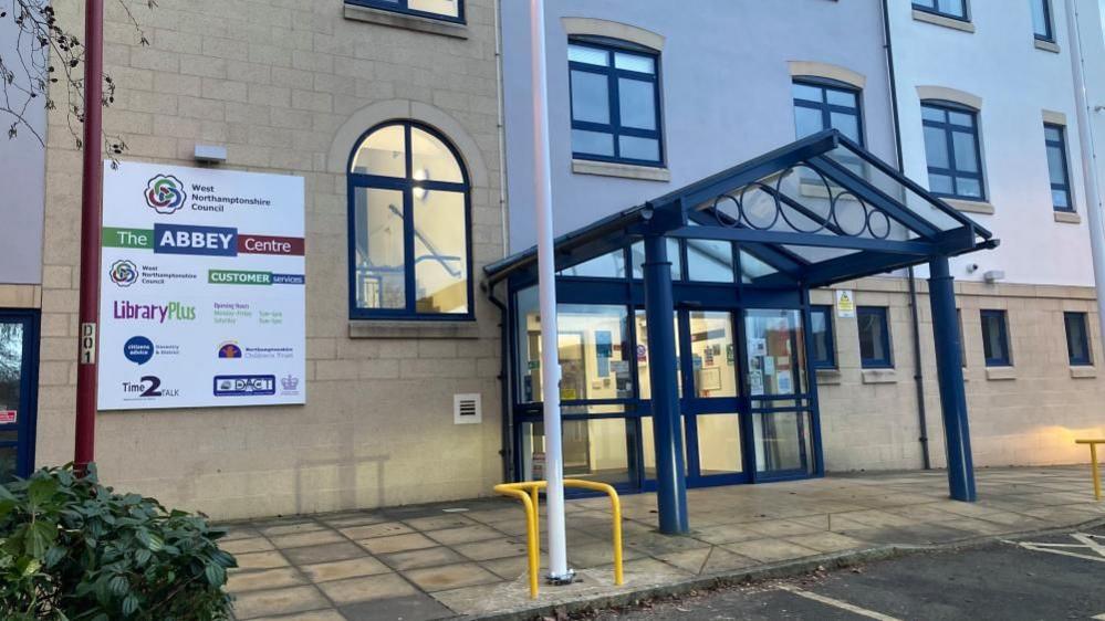 Three-storey building in light coloured brick with glass entrance doors with blue frames. There is a paved area outside and parking spaces.  A sign says "The Abbey Centre" with the names of organisations located in the building, including West Northamptonshire Council, Time2Talk and DACT.