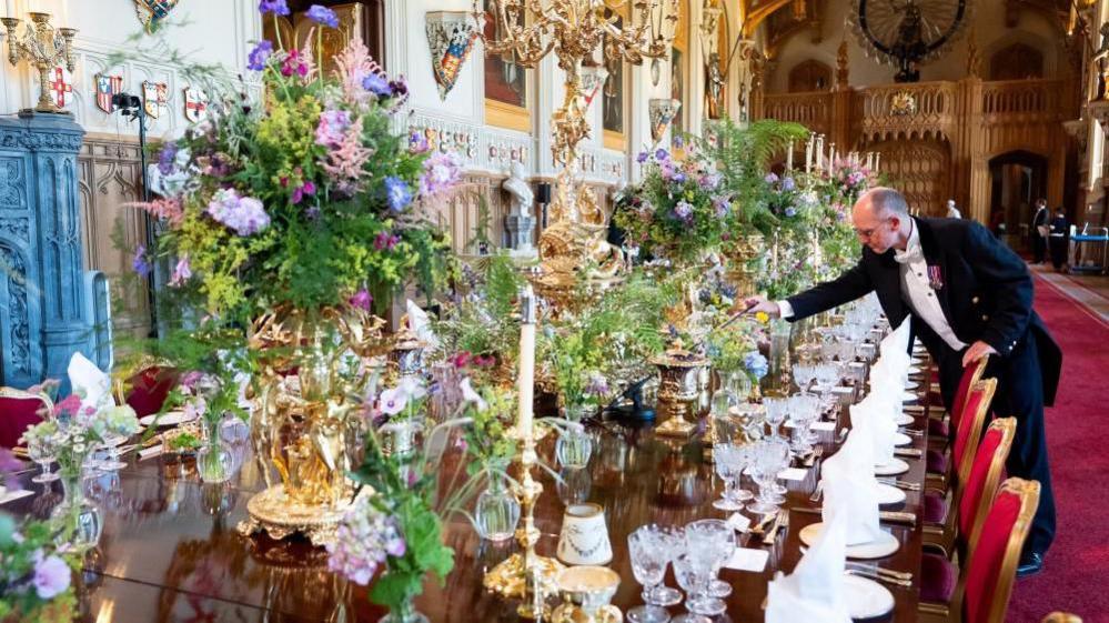 A member of staff is making the final touches to a long table set with elaborate bouquets of flowers in gold vases and table-wear in Windsor Castle.