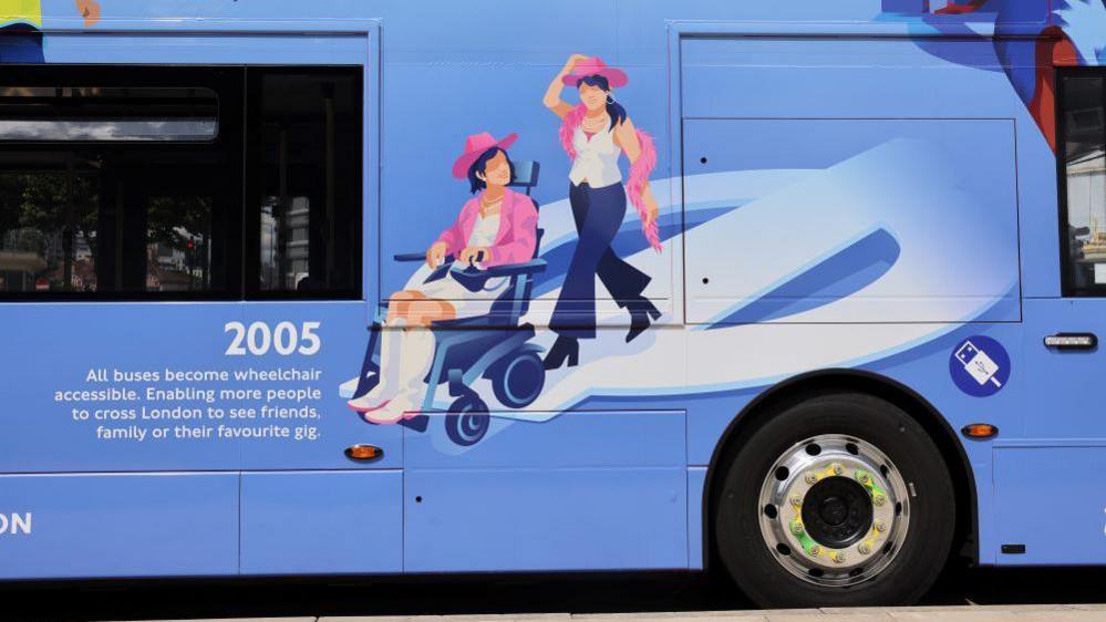 A close-up of the side of a sky-blue coloured bus. The focus of the image is the cartoon drawing of two women, one of whom is in a dark blue wheelchair. The woman in a wheelchair is wearing white cowboy boots, a white dress, a bright pink jacket, and a pink cowboy hat, and has short black hair. The other woman is walking just behind her, has long black hair, and is wearing black boots, blue jeans, a white top, a pink cowboy hat, and a pink feather boa. A white Transport for London roundel is integrated into the design to represent a wheelchair ramp. On the left of the design, just below one of the bus windows, there is writing reading: "2005 All buses become wheelchair accessible. Enabling more people to cross London to see friends, family or their favourite gig.". One of the wheels of the bus is beneath the design on the right hand side.