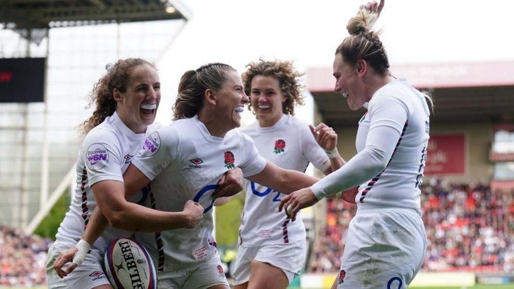 Four England player in an all white kit celebrate a try. Behind them is two stands filled with fans.