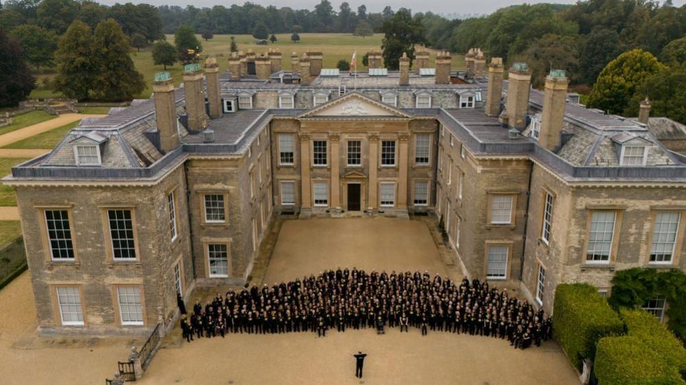 Two-storey stone stately home with courtyard. A semicircle of singers is standing in rows in front of the house, with a conductor facing them.