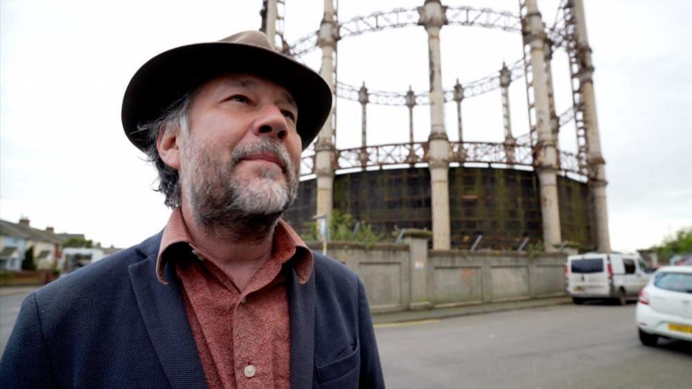 Joe Mackintosh is bearded and wearing a trilby hat. He is also wearing a blue jacket with a rust-brown shirt beneath. He is standing near the Great Yarmouth gasometer, which sits within a walled compound. There are vehicles parked on the right of the image, and a terrace of houses visible on the left of the image.