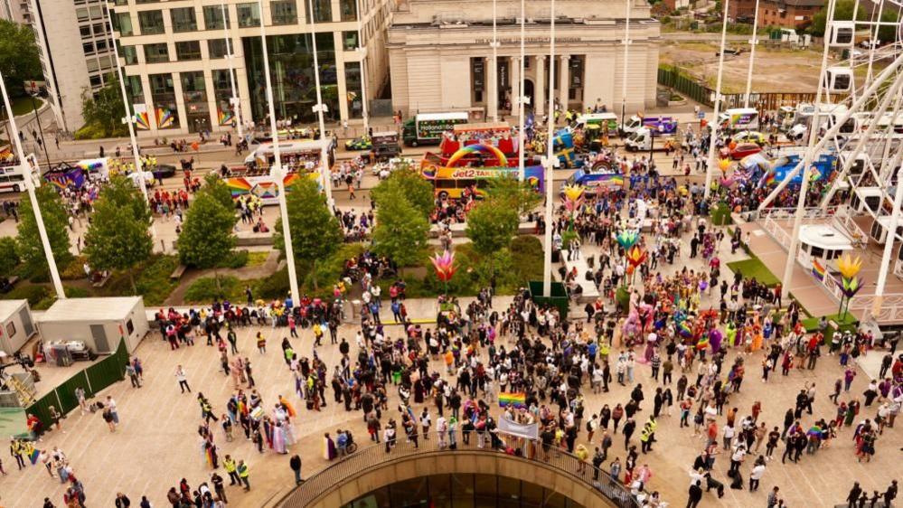 An aerial shot looking down on a busy square in a city during its Pride festival. People are gathered holding Pride flags and dressed in costumes and bright colours. There are a number of buses and floats behind them that are part of the parade. The edge of a big wheel is on the right. 