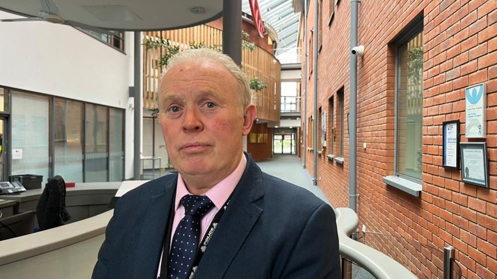 Andy Guy, standing in the atrium of Norfolk Police headquarters. The building has a red brick wall to the right of the image, featuring grey windows and a couple of certificates in frames mounted on the wall. There is a reception counter to the left of the picture, and architectural features including railings, wood cladding and glazing to the left and centre of the image.