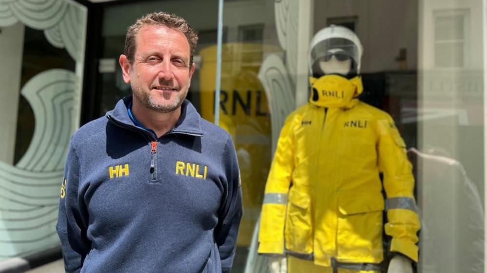 RNLI crew member, Mike Edward stands in front of the shop housing the exhibition, with a mannequin wearing RNLI uniform in the shop window behind him