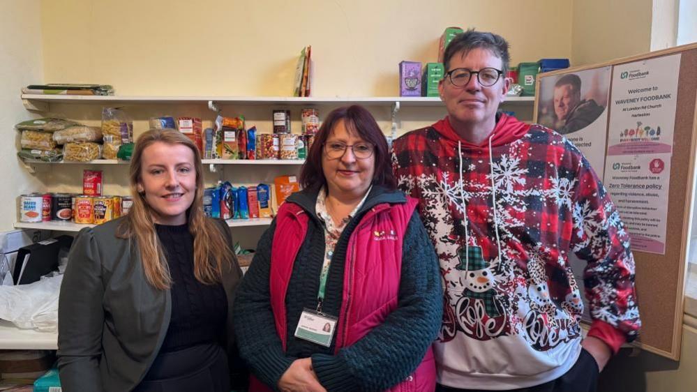 Laura Rawnsley, on the left, has long blonde hair. She is wearing a black jersey and grey jacket. Next to her is Jemma Jackson, the operations manager of the Waveney Food Bank, wearing a blue knitted jersey and pink gilet. She has burgundy-coloured hair. Matthew Scade, the food bank project manager, is wearing a brightly patterned Christmas-them jersey. He has medium length dark hair and wears glasses.