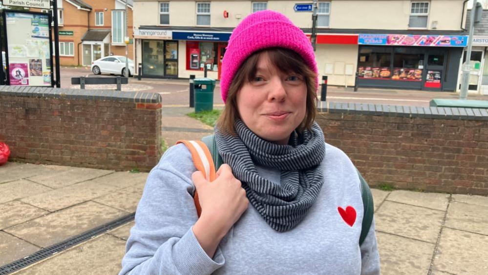 Joanne Ollier, standing outside a row of shops that are behind her. She is wearing a bright pink hat, a blue and black scarf, a blue jumper, with a red heart to the right. She has one arm on her shoulder, holding on to a bag strap that is cream and orange.