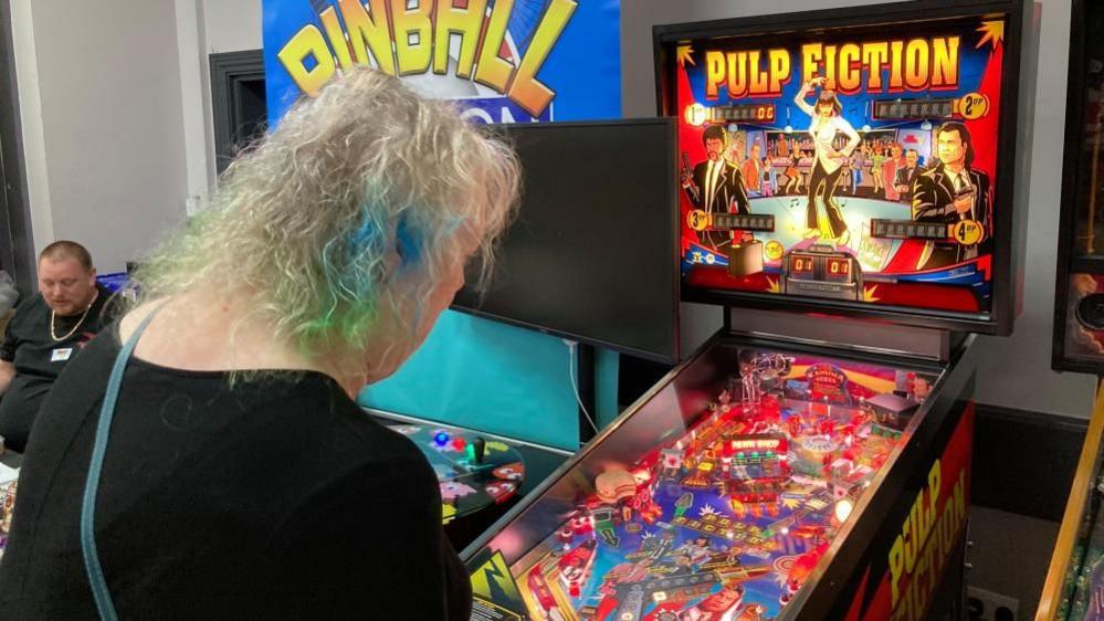 Woman with long coloured hair plays on a machine with Pulp Fiction artwork, including a depiction of John Travolta playing a guitar