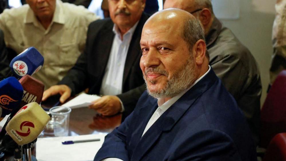 A photograph of Khalil al-Hayya. He is sat at a news conference and three other men can be seen in the background behind him. He is wearing a dark blue suit.