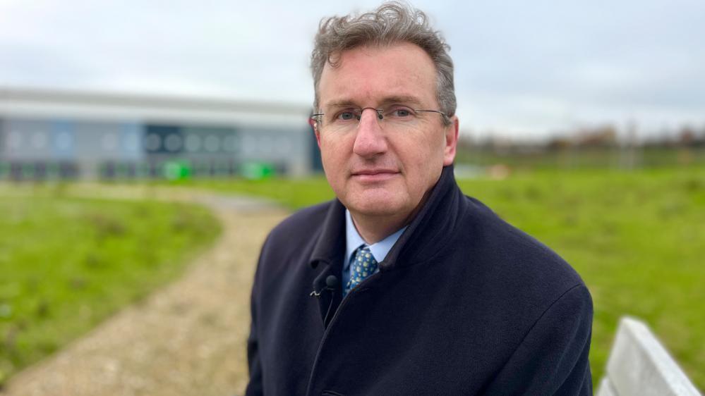 A man is sitting on a bench, wearing glasses and appearing with greying hair. There is a landscaped background with a warehouse in the background. The man is wearing a dark suit and tie.