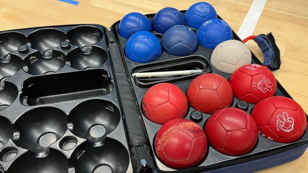 A set of Boccia balls in a black case. The case has holes for the balls to sit in and six red balls and six blue balls fill one side, divided by a white ball and a white measuring stick.