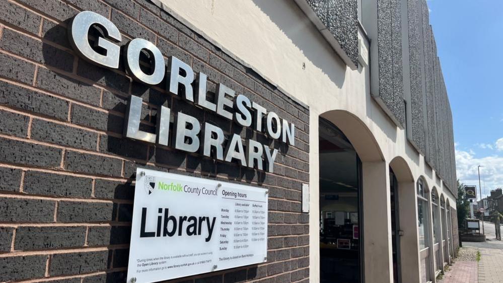 Chrome signage stating "Gorleston Library" and the library opening hours on a tablet below, bearing the logo and name of Norfolk County Council. The building is a 1970s design, featuring brown bricks, concrete pillars and arches, several infilled with aluminium-framed windows, and flint-faced concrete panels on the upper storey.