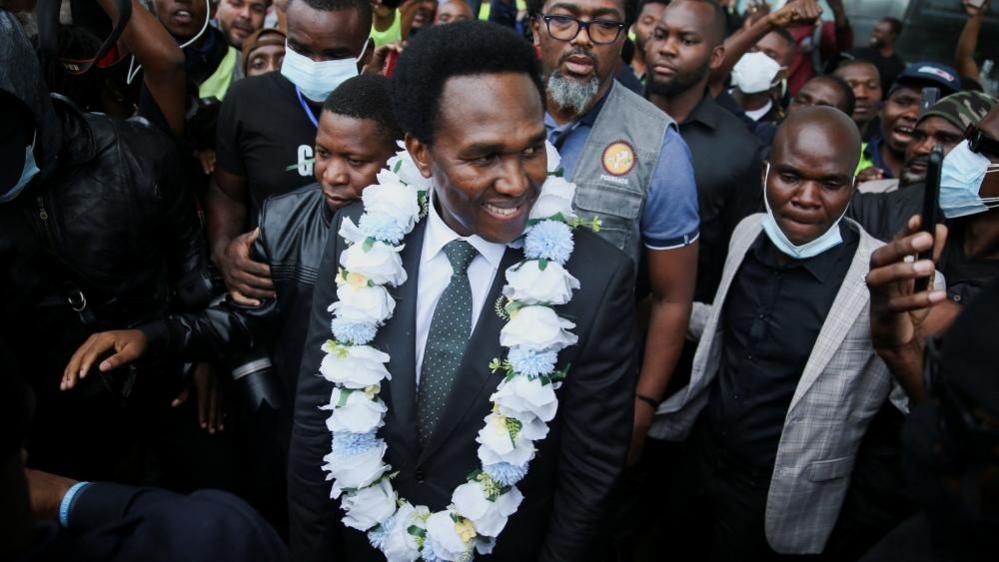 Venancio Mondlane comes outside the terminal building at the Maputo International Airport wearing a white garland around his next and surrounded by journalists and supporters.