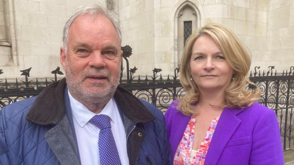 David Fraser and Liz Furber in front of a court building. David is wearing a shirt and tie with a blue coat, and Liz is wearing a multicoloured top with a purple jacket.