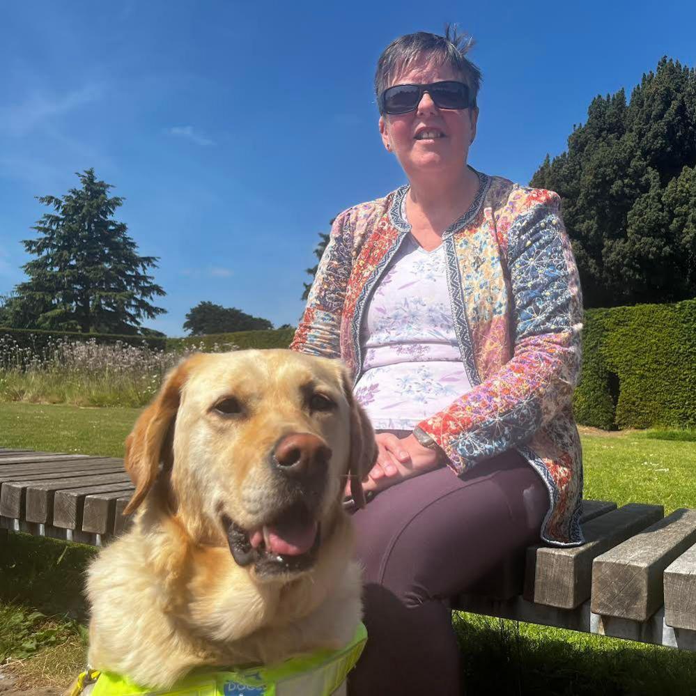 Sylvia Paton sits on a summers day on a wooden bench in the sun with blue sky. She is wearing sunglasses as bright light affects her eye condition. She has her blonde Labrador dog, Kate, in the foreground.