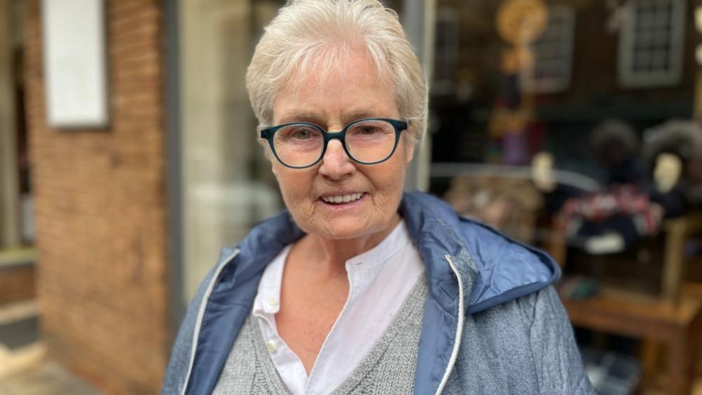 A picture of Maggie Clark, who is wearing glasses, a shirt, vest and jacket.