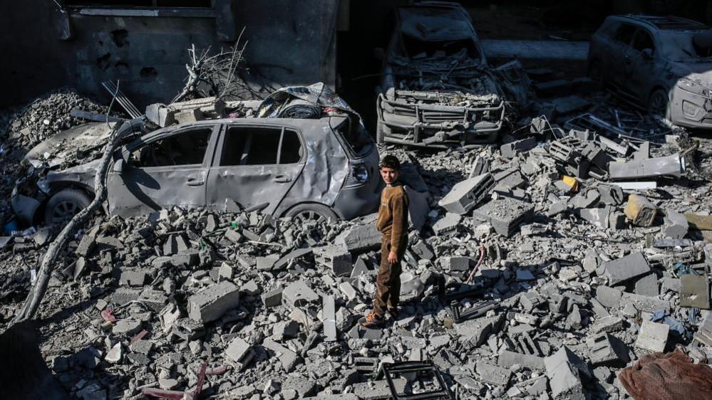 A pre-teen boy stands amongst grey rubble and burned out cars, squinting up towards the camera