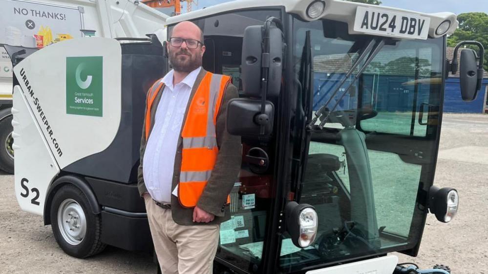 Councillor Paul Wells in a high visibility orange tabard, wearing a white shirt, jacket and chinos. He has a full beard and is bald, and wears glasses. He is standing next to a Great Yarmouth Borough Services street sweeping machine.