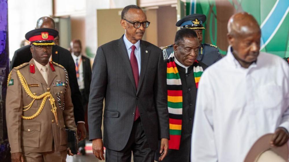 Rwandan President Paul Kagame, wearing a black striped suit and red tie, walks among a group of men