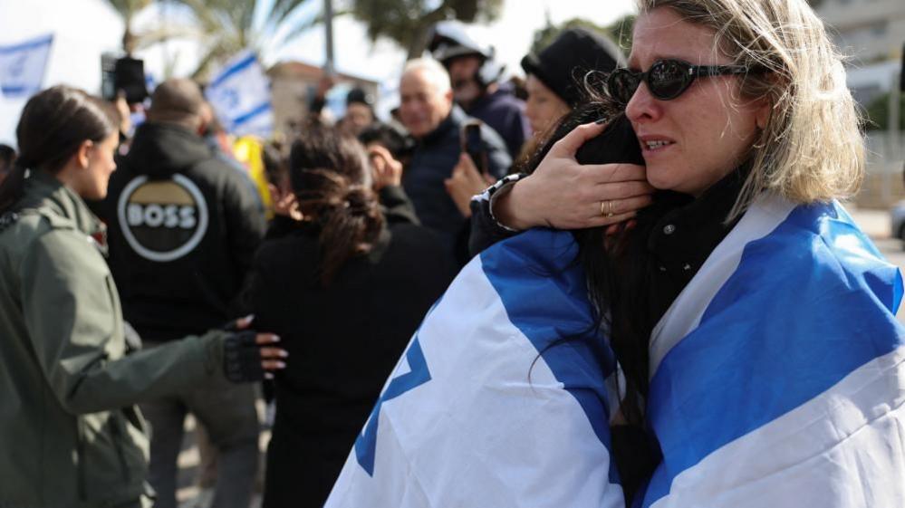 A distressed-looking woman draped in an Israeli flag holds another person (20/02/25)