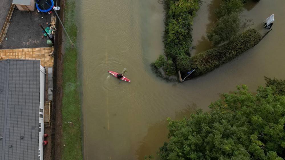 A red canoe is turning a corner on floodwater with hedges either side. A grey-roofed building is visible to the left. A cafe sign can just be seen to the right.