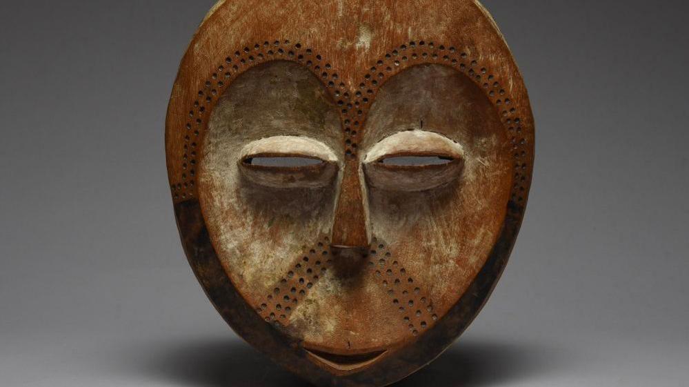 A mask depicting a stylised version of a human face - there is a small smile, narrow eyes and dots pressed into surface which go in a horse-show shape where eyebrows would be and are all under the nose.