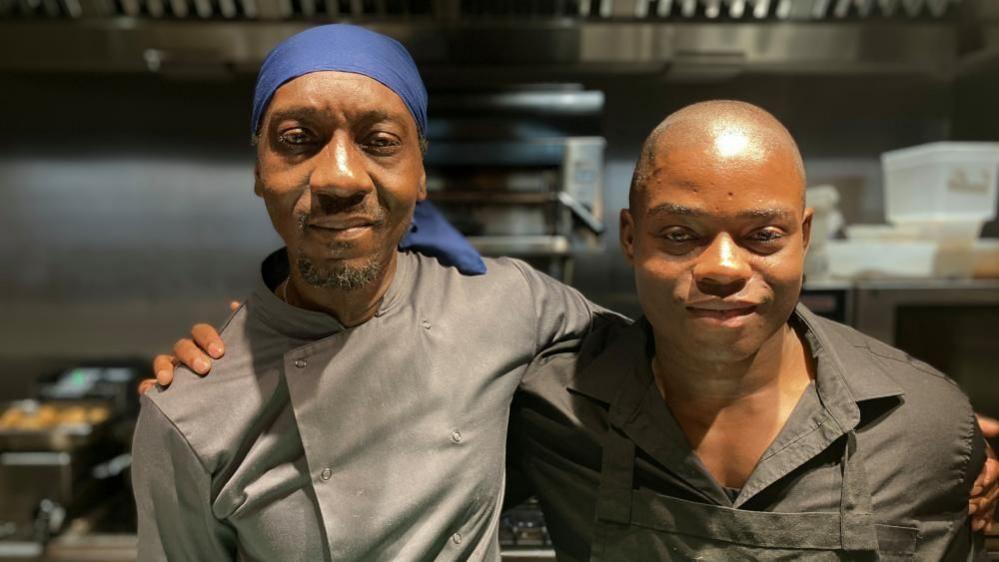 Two men wearing chef's clothes stand with their arms around each other in an industrial kitchen. 