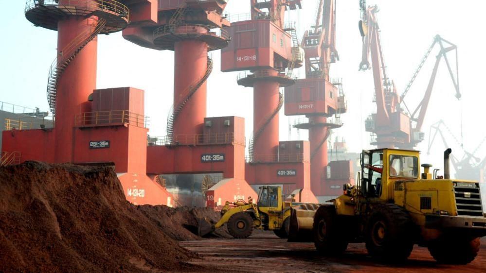 Two yellow trucks move heaps of soil containing rare earth elements at a port in China. At least five red cranes in the background tower above the trucks.