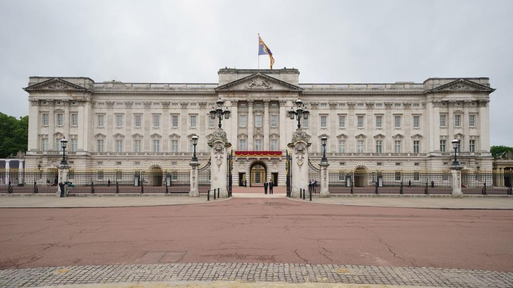 A frontal view of Buckingham Palace. The only people in the shot are two police officers standing just inside the palace's open gates. 