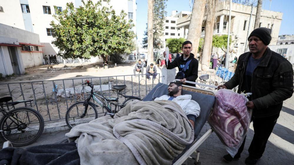 An injured person lies in a stretcher, which is being wheeled by two men. Two bicycles are parked by a metal fence and there are buildings around. 